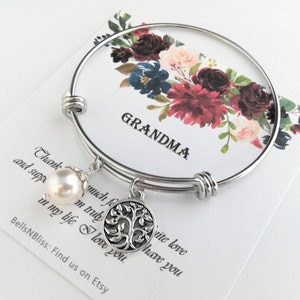 May include: A silver-toned bangle bracelet with a dangling pearl and a tree of life charm. The bracelet is displayed on a card with the word "GRANDMA" and a floral design in burgundy, navy, and blush tones. The card also has text.