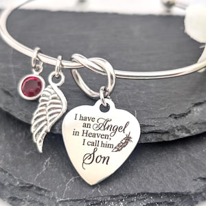 May include: Silver-toned bangle bracelet with three charms. One charm is a red gemstone, another is a feather, and the third is a heart-shaped pendant with the inscription "I have an Angel in Heaven, I call him Son."