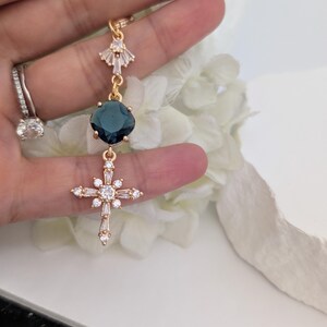 May include: A gold-toned pendant featuring a fan-shaped top element with clear crystals, a central square dark blue gemstone, and a lower cross-shaped charm adorned with multiple clear crystals. The piece is suspended from a delicate gold-toned chain.