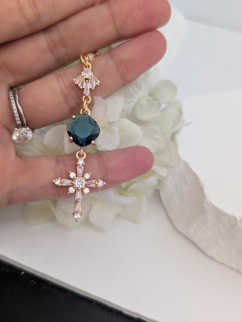May include: A gold-toned pendant featuring a fan-shaped top element with clear crystals, a central square dark blue gemstone, and a lower cross-shaped charm adorned with multiple clear crystals. The piece is suspended from a delicate gold-toned chain.