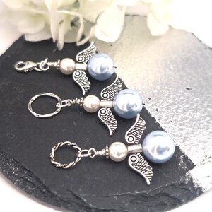 May include: Three angel-themed keychains with silver wings, pearl beads, and light blue pearl accents. One keychain has a clasp, and two have key rings. The keychains are arranged on a black and silver surface, with white flowers in the background.