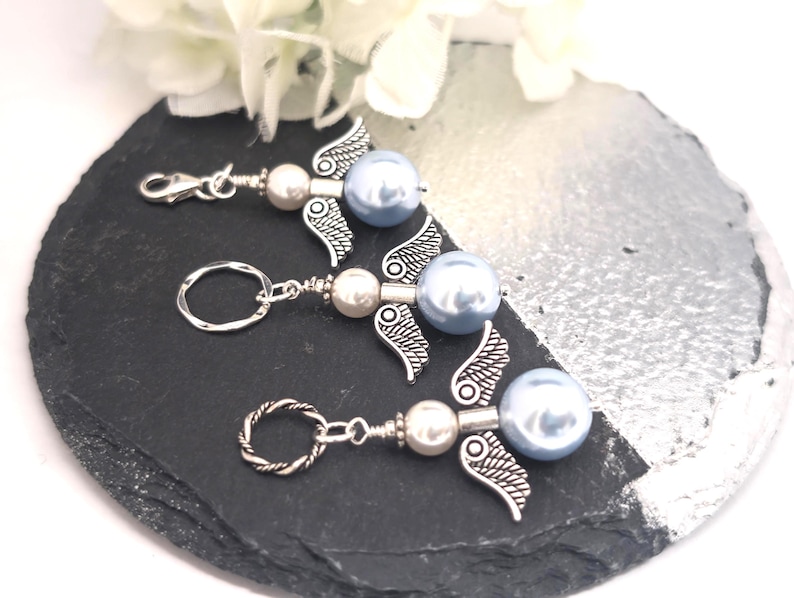 May include: Three angel-themed keychains with silver wings, pearl beads, and light blue pearl accents. One keychain has a clasp, and two have key rings. The keychains are arranged on a black and silver surface, with white flowers in the background.