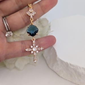 May include: A gold-toned pendant featuring a fan-shaped top element with clear crystals, a central round dark blue faceted stone, and a lower cross-shaped charm adorned with multiple clear baguette and round crystals in a starburst arrangement.