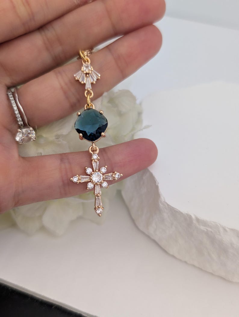 May include: A gold-toned pendant featuring a fan-shaped top element with clear crystals, a central round dark blue faceted stone, and a lower cross-shaped charm adorned with multiple clear baguette and round crystals in a starburst arrangement.