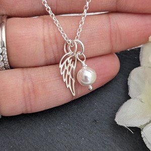 May include: A delicate silver necklace featuring a small, openwork wing charm and a dangling white pearl. The chain is fine and the pendant is centered. The necklace is shown against a soft background.