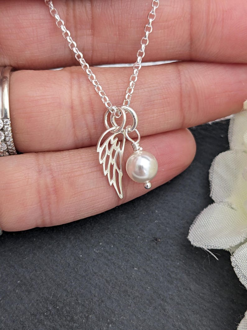 May include: A delicate silver necklace featuring a small, openwork wing charm and a dangling white pearl. The chain is fine and the pendant is centered. The necklace is shown against a soft background.
