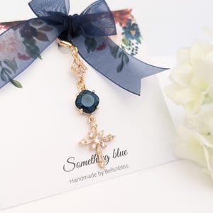 May include: A gold-toned charm featuring a dark blue square crystal and a cross-shaped pendant adorned with clear stones. The charm is attached to a white display card with a navy blue organza ribbon, featuring the text Something blue Handmade by Bellsnbliss.