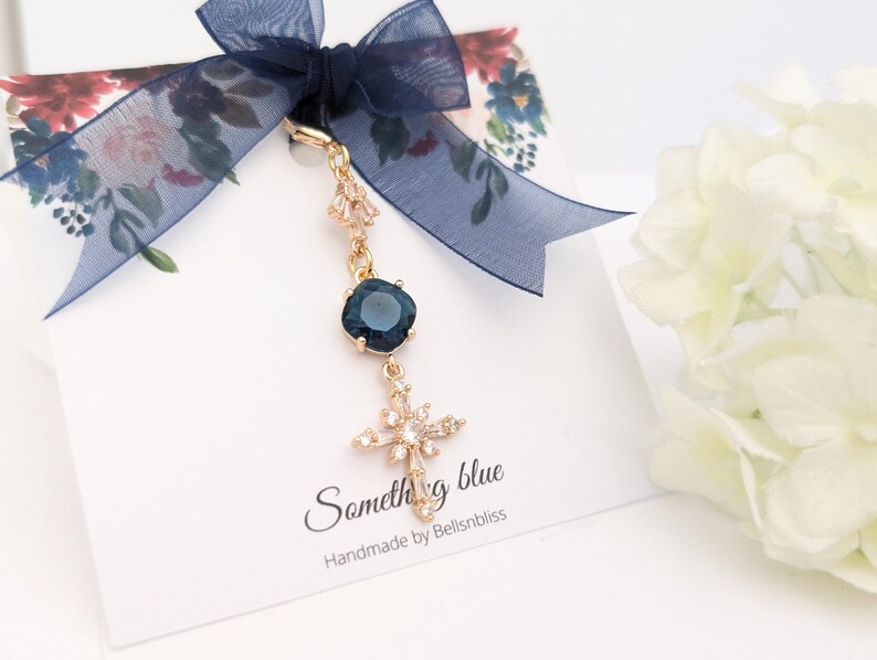 May include: A gold-toned charm featuring a dark blue square crystal and a cross-shaped pendant adorned with clear stones. The charm is attached to a white display card with a navy blue organza ribbon, featuring the text Something blue Handmade by Bellsnbliss.