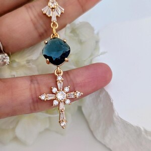 May include: A gold-toned drop earring featuring a fan-shaped top accent with clear crystals, a central dark blue faceted gemstone, and a lower cross-shaped charm adorned with clear baguette and round crystals set in a gold-colored metal frame.