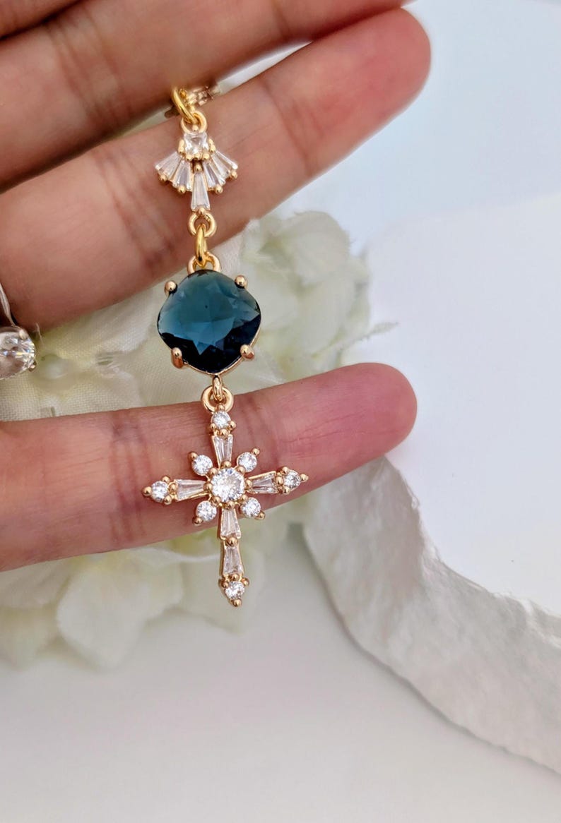 May include: A gold-toned drop earring featuring a fan-shaped top accent with clear crystals, a central dark blue faceted gemstone, and a lower cross-shaped charm adorned with clear baguette and round crystals set in a gold-colored metal frame.