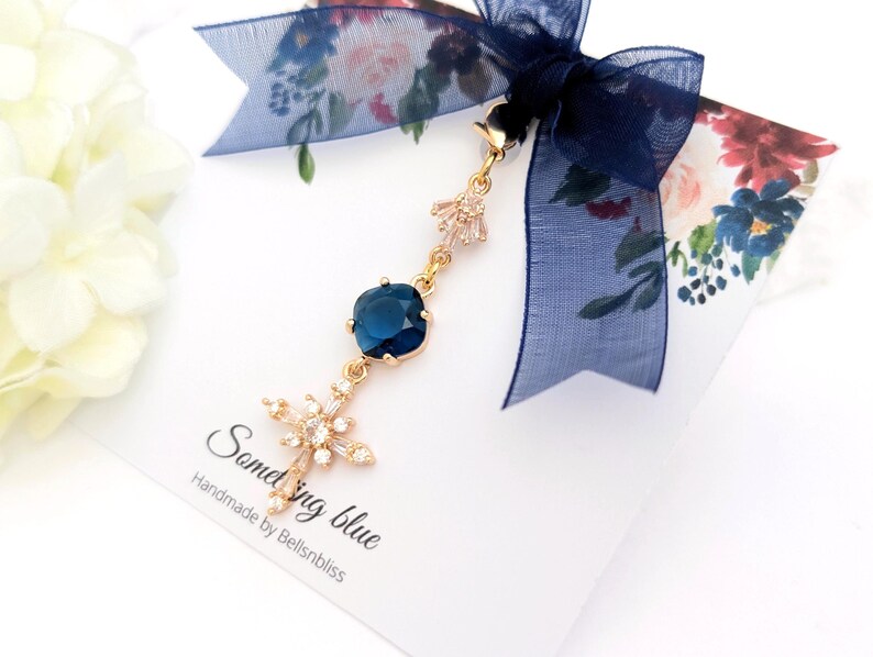 May include: A gold-toned charm featuring a deep blue square-cut crystal and a star-shaped crystal pendant, suspended from a dark blue sheer ribbon bow. The charm is attached to a white display card with the text Something blue Handmade by Bellsnbliss.