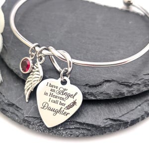 Op de afbeelding: Zilverkleurige armband met een hartvormige bedel. De bedel is gegraveerd met de woorden "Ik heb een engel in de hemel, ik noem haar dochter". Het heeft ook een rode edelsteen en een bladbedel.