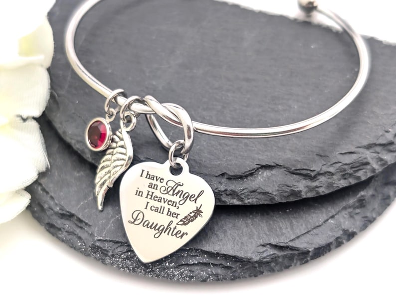 Op de afbeelding: Zilverkleurige armband met een hartvormige bedel. De bedel is gegraveerd met de woorden "Ik heb een engel in de hemel, ik noem haar dochter". Het heeft ook een rode edelsteen en een bladbedel.