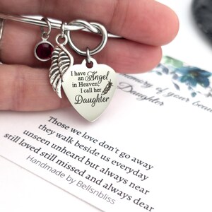 Op de afbeelding: Zilverkleurige armband met een hartvormige bedel met de inscriptie "I have an Angel in Heaven, I call her Daughter." Inclusief een rode edelsteen en een veervormige bedel. Een kaart met een gedicht is zichtbaar.