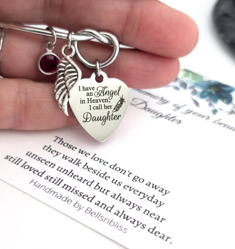 Op de afbeelding: Zilverkleurige armband met een hartvormige bedel met de inscriptie "I have an Angel in Heaven, I call her Daughter." Inclusief een rode edelsteen en een veervormige bedel. Een kaart met een gedicht is zichtbaar.