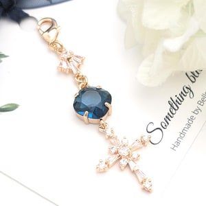 May include: A gold-toned metal charm featuring a lobster clasp, a small clear crystal accent, a central square-cut dark blue gemstone in a prong setting, and a cross-shaped pendant adorned with multiple small clear crystals.