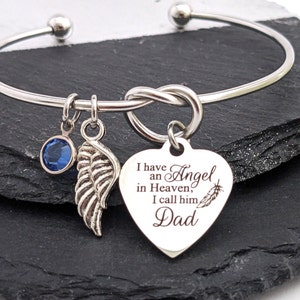 May include: A silver-toned bangle bracelet with three charms. One charm is a blue gemstone, another is a wing, and the third is a heart-shaped pendant with the text "I have an Angel in Heaven, I call him Dad".
