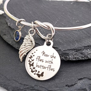 May include: Silver bangle bracelet with a charm that reads "Now she flies with butterflies". The charm is surrounded by a circle of small butterfly charms. The bracelet also has a small blue crystal charm and a silver wing charm.