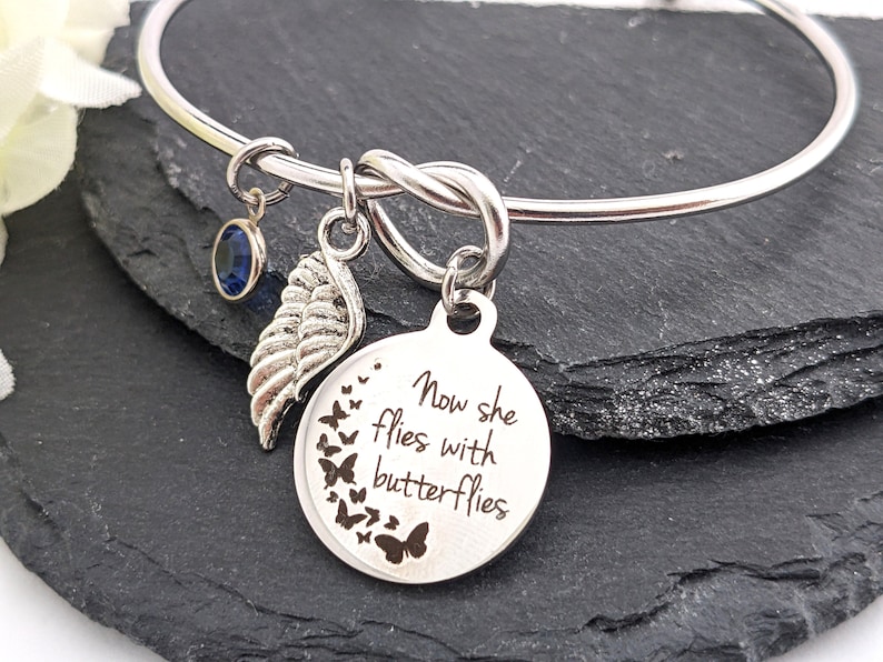 May include: Silver bangle bracelet with a charm that reads "Now she flies with butterflies". The charm is surrounded by a circle of small butterfly charms. The bracelet also has a small blue crystal charm and a silver wing charm.