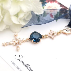 May include: A gold-toned metal charm featuring a central square dark blue crystal, a cross-shaped pendant adorned with clear baguette and round crystals, and a smaller crystal-encrusted accent piece, all attached to a lobster clasp with a navy ribbon.