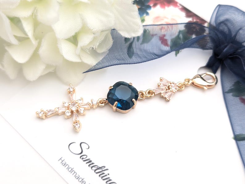 May include: A gold-toned metal charm featuring a central square dark blue crystal, a cross-shaped pendant adorned with clear baguette and round crystals, and a smaller crystal-encrusted accent piece, all attached to a lobster clasp with a navy ribbon.