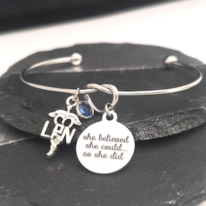 May include: A silver-colored bangle bracelet with three charms. One charm is a caduceus symbol with the letters LPN. Another is a small blue gemstone. The third is a round charm with the engraved text: "she believed she could... so she did."