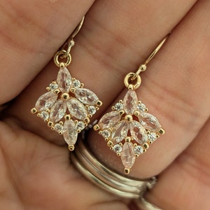 May include: A pair of gold earrings with a diamond-shaped design. The earrings are adorned with clear, marquise-shaped stones.