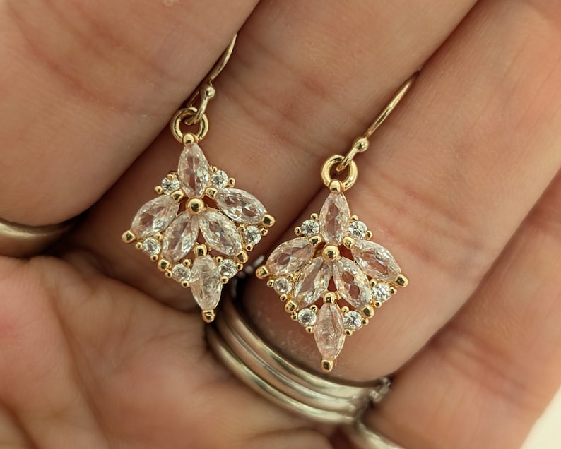May include: A pair of gold earrings with a diamond-shaped design. The earrings are adorned with clear, marquise-shaped stones.