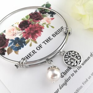 May include: A silver bangle bracelet with a tree of life charm and a dangling pearl. The bracelet is displayed on a card with floral accents and the text "MOTHER OF THE BRIDE".