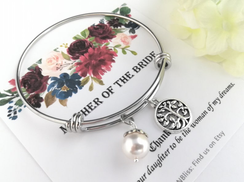 May include: A silver bangle bracelet with a tree of life charm and a dangling pearl. The bracelet is displayed on a card with floral accents and the text "MOTHER OF THE BRIDE".