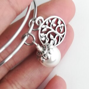May include: Silver-toned bangle bracelet with two charms. One charm is a round, openwork design of a tree with leaves. The other is a white, pearlescent bead with a decorative silver cap.