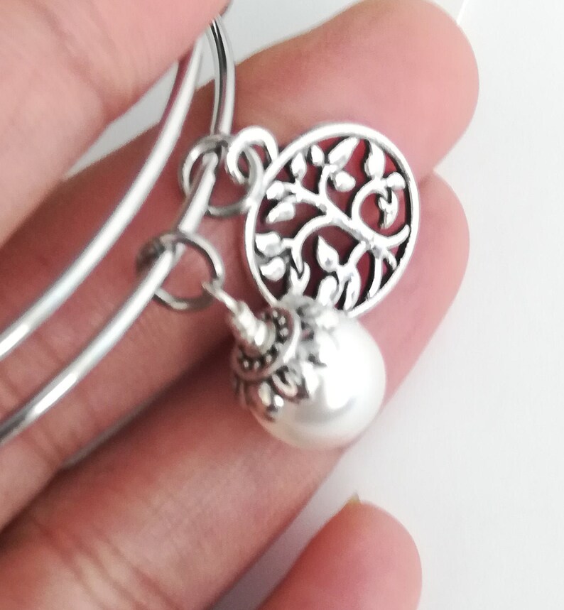 May include: Silver-toned bangle bracelet with two charms. One charm is a round, openwork design of a tree with leaves. The other is a white, pearlescent bead with a decorative silver cap.