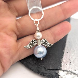 May include: A silver angel charm with a light blue pearl body, a white pearl head, and silver wings. The charm is attached to a sheer white ribbon. The angel is a decorative item.