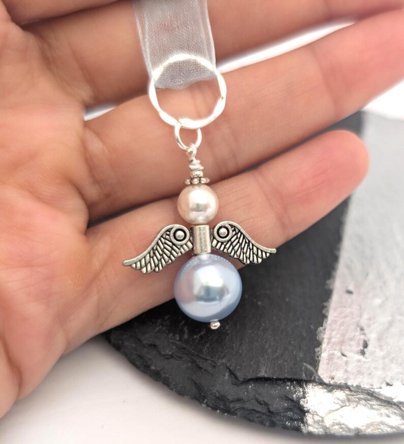 May include: A silver angel charm with a light blue pearl body, a white pearl head, and silver wings. The charm is attached to a sheer white ribbon. The angel is a decorative item.