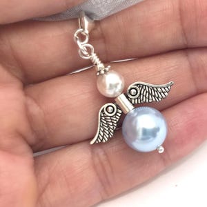 May include: A silver-toned angel charm with pearl accents and delicate wings. The charm features a light blue pearl body and a white pearl head, suspended from a silver chain and a sheer ribbon.