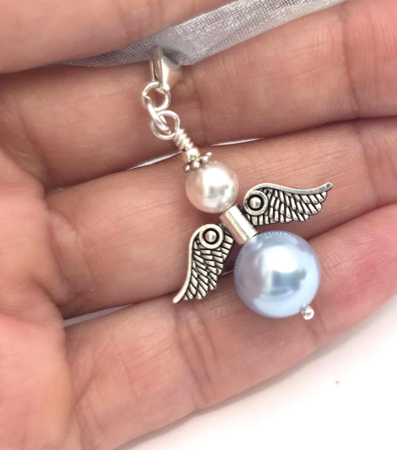 May include: A silver-toned angel charm with pearl accents and delicate wings. The charm features a light blue pearl body and a white pearl head, suspended from a silver chain and a sheer ribbon.