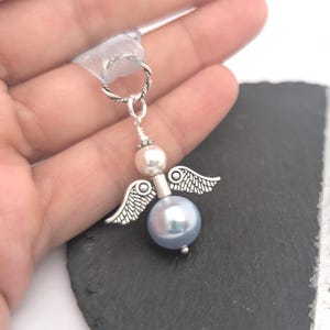 May include: A silver-toned angel charm with a light blue pearl body, a white pearl head, and small silver wings. The charm is suspended from a silver-toned loop with a white ribbon. The angel is held in a hand.