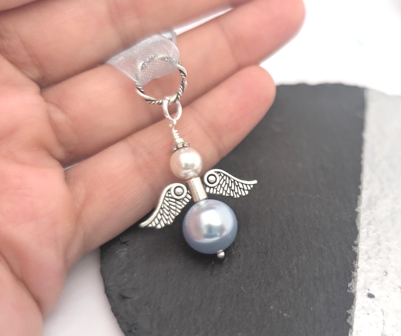May include: A silver-toned angel charm with a light blue pearl body, a white pearl head, and small silver wings. The charm is suspended from a silver-toned loop with a white ribbon. The angel is held in a hand.