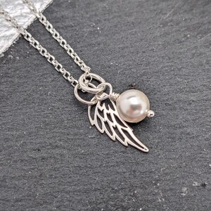 May include: A silver necklace with a delicate chain. The pendant features a pearl and a wing charm. The wing charm is intricately detailed. The necklace is displayed on a dark gray surface.