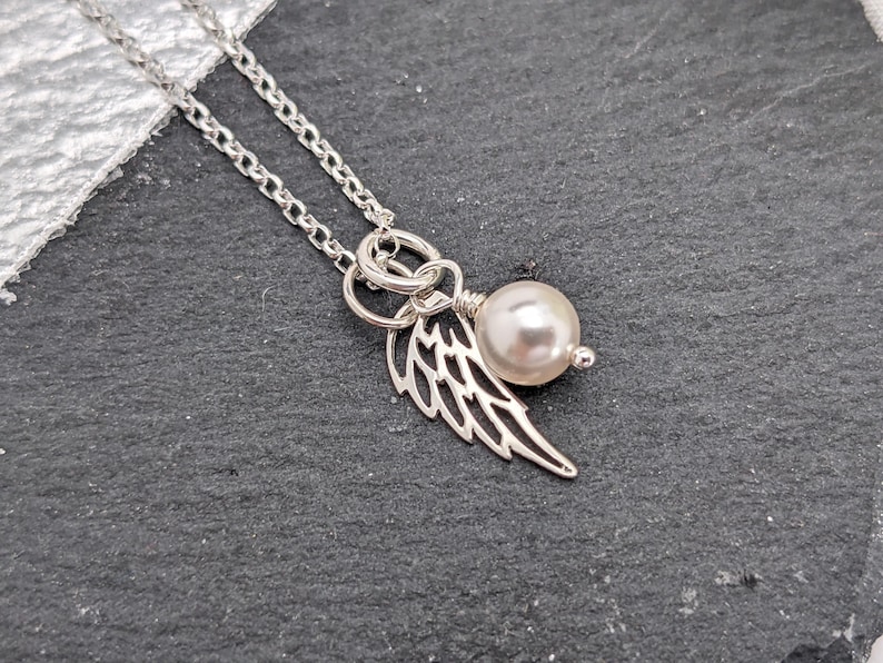 May include: A silver necklace with a delicate chain. The pendant features a pearl and a wing charm. The wing charm is intricately detailed. The necklace is displayed on a dark gray surface.