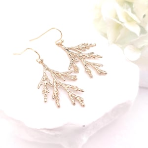 May include: A pair of gold-toned drop earrings featuring a textured, branch-like design that mimics the appearance of a small conifer twig. The earrings are suspended from simple gold-toned ear wires, showcasing a detailed, organic, and metallic finish.