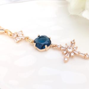 May include: A gold-toned charm featuring a lobster clasp, a small fan-shaped clear crystal accent, a central square-cut deep blue crystal in a prong setting, and a dangling cross-shaped pendant adorned with multiple small clear baguette crystals.