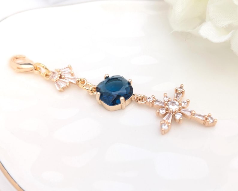 May include: A gold-toned charm featuring a lobster clasp, a small fan-shaped clear crystal accent, a central square-cut deep blue crystal in a prong setting, and a dangling cross-shaped pendant adorned with multiple small clear baguette crystals.