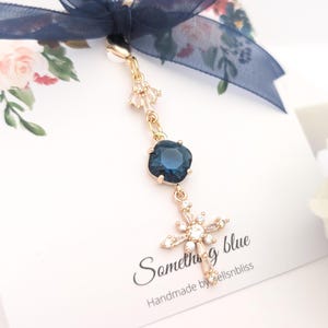 May include: A gold-toned charm featuring a dark blue square crystal, a small clear crystal accent, and a clear crystal cross pendant. The charm is attached to a navy blue ribbon bow on a white card with the text Something blue and Handmade by bellsnbliss.