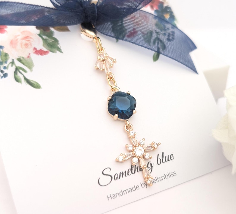 May include: A gold-toned charm featuring a dark blue square crystal, a small clear crystal accent, and a clear crystal cross pendant. The charm is attached to a navy blue ribbon bow on a white card with the text Something blue and Handmade by bellsnbliss.