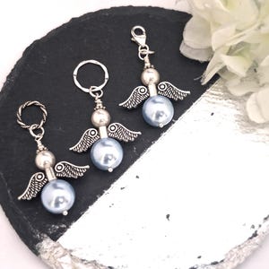 May include: Three angel-themed charms with silver-tone wings and accents. Each charm features a light blue pearl-like bead as the body. One charm has a keyring, one has a lobster clasp, and one has a twisted ring. The charms are displayed on a black and silver surface.
