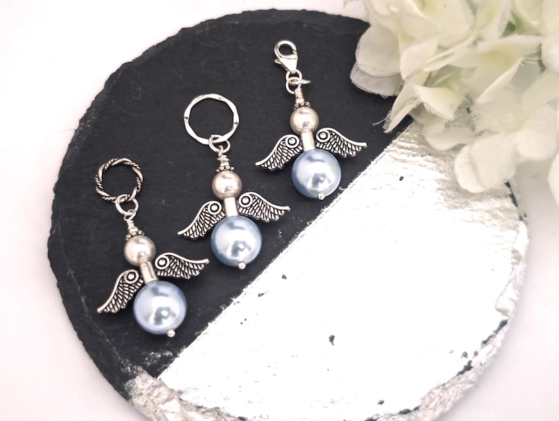 May include: Three angel-themed charms with silver-tone wings and accents. Each charm features a light blue pearl-like bead as the body. One charm has a keyring, one has a lobster clasp, and one has a twisted ring. The charms are displayed on a black and silver surface.
