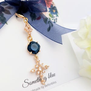 May include: A gold-toned charm featuring a dark blue square crystal and a cross pendant adorned with clear stones, suspended from a navy blue sheer ribbon. The charm is attached to a white display card with the text Something blue and Bellsnbliss.