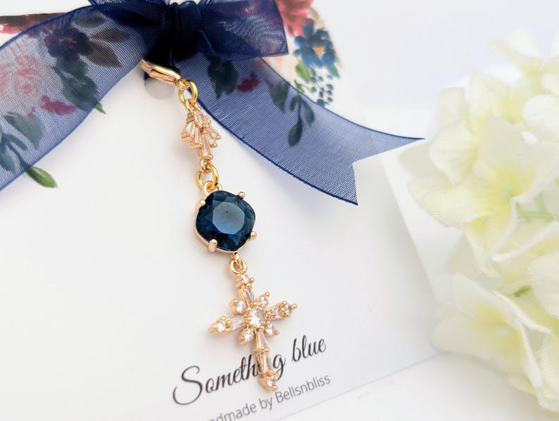 May include: A gold-toned charm featuring a dark blue square crystal and a cross pendant adorned with clear stones, suspended from a navy blue sheer ribbon. The charm is attached to a white display card with the text Something blue and Bellsnbliss.