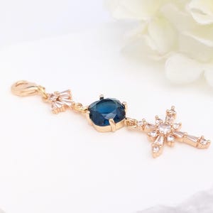 May include: A gold-toned charm featuring a lobster clasp, a small cluster of clear crystals, a central square-cut deep blue gemstone in a prong setting, and a dangling cross pendant embellished with multiple clear, baguette-cut crystals.